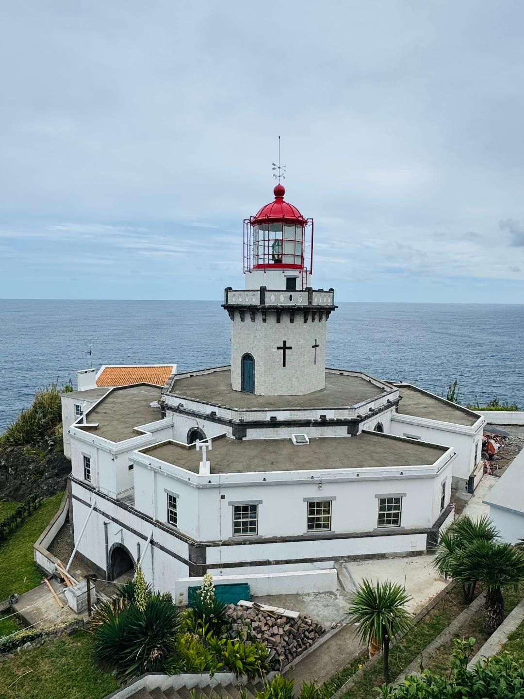 A white, hexagonal lighthouse with a red-topped lantern room on a cliff overlooking the blue ocean under a cloudy sky.