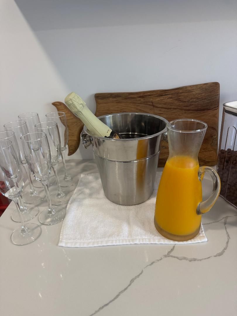 A silver ice bucket with a champagne bottle, a pitcher of orange juice, and several glass flutes on a white counter.