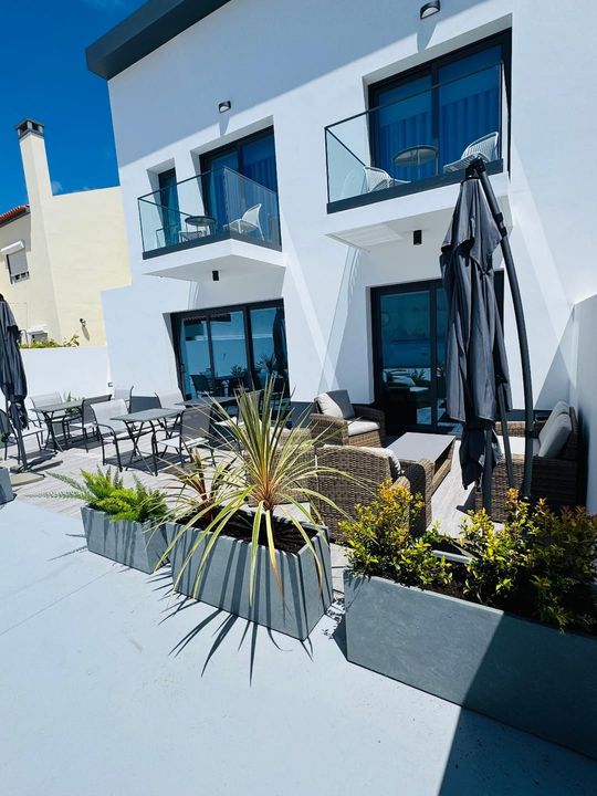 A modern white two-story building with balconies, an outdoor patio, seating, and potted greenery on a bright sunny day.