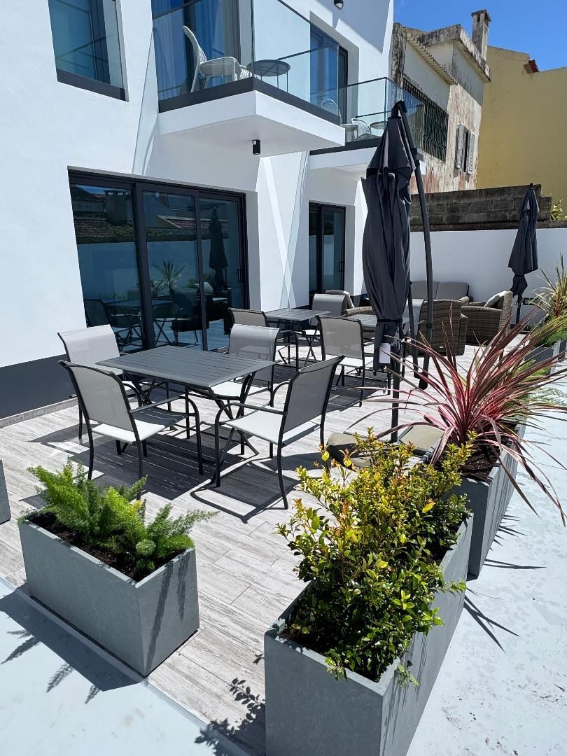 Outdoor patio with a dining table and chairs, potted plants, and gray umbrellas next to a modern white building.