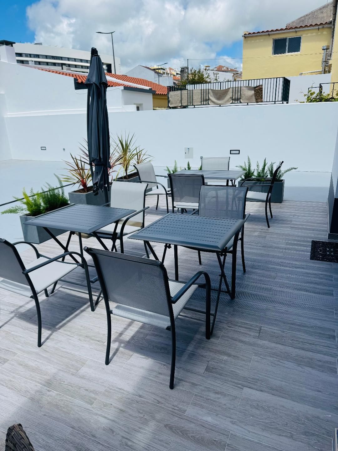 A rooftop terrace with gray metal tables and chairs, potted plants, and a closed patio umbrella against a white wall.
