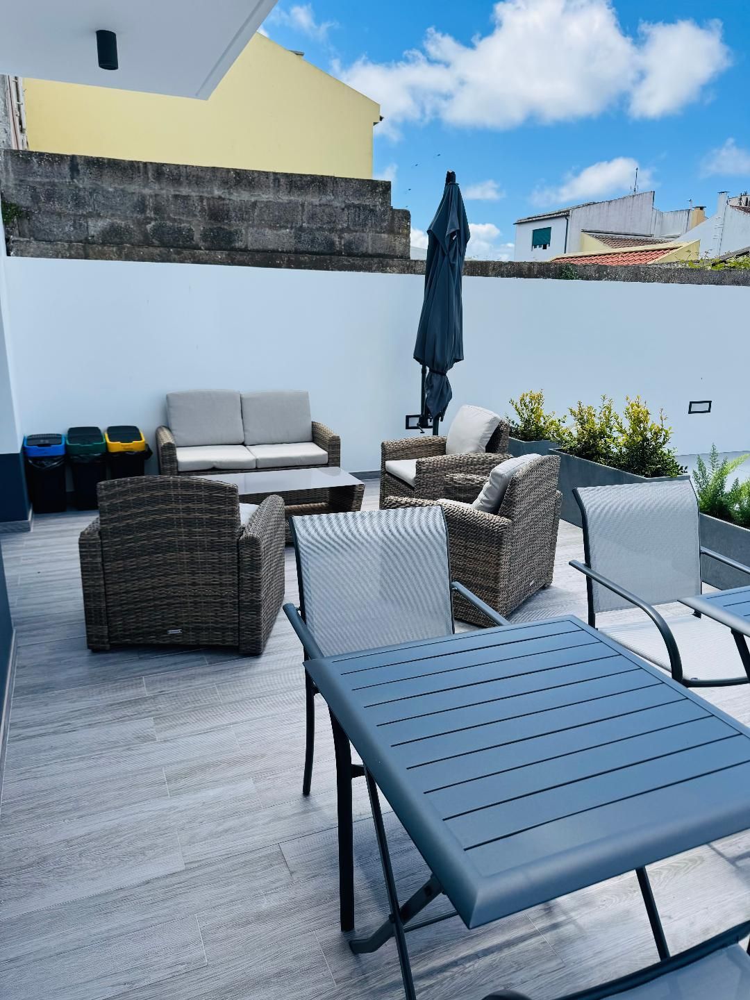 A patio terrace furnished with wicker and metal seating, a small table, and three waste bins against a white wall.