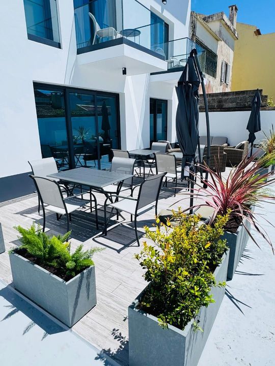 Outdoor patio with modern tables, chairs, and potted plants under a clear blue sky by a white building.