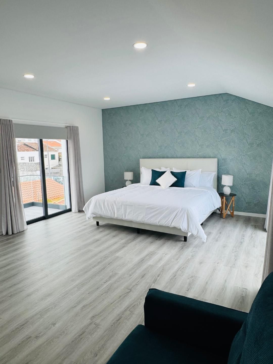 A bright bedroom with a bed against a teal textured wall, light wood flooring, floor-to-ceiling windows, and a blue chair.