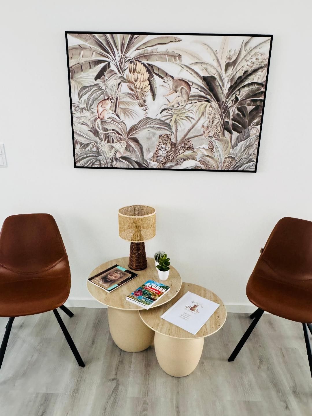 Two brown chairs flank a nesting table with a lamp, a small plant, and two magazines under a framed tropical wall art.