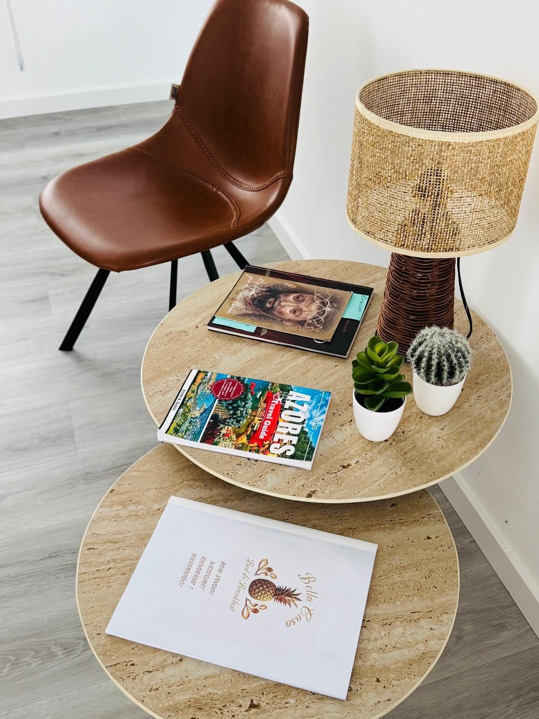 A brown leather chair beside stacked round light-wood tables with a woven lamp, two small plants, and books.