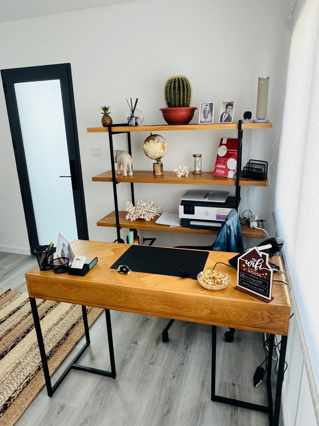 A modern wooden desk with a black frame and a matching three-tier shelving unit against a white wall in a home office.