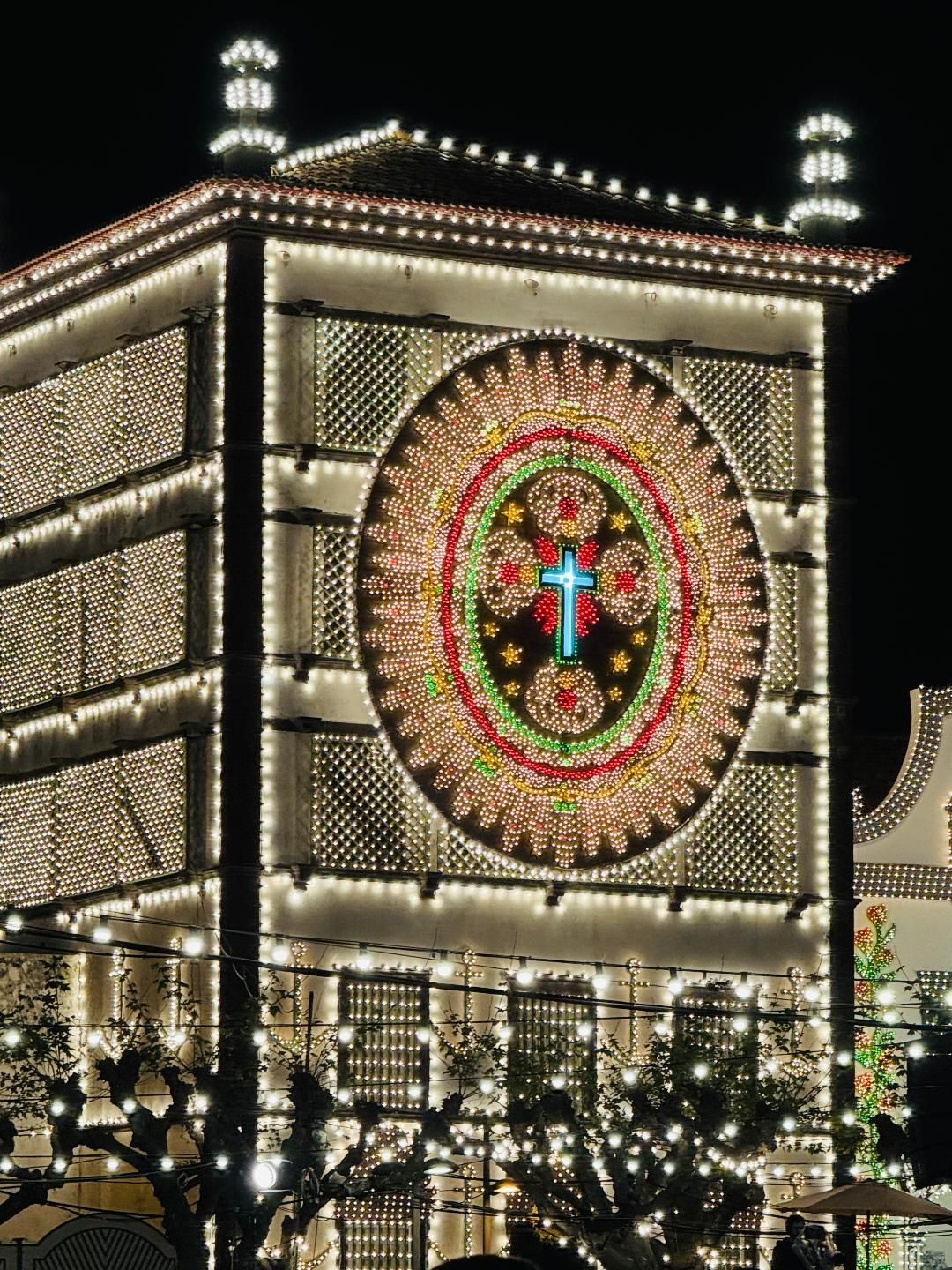 A tall building at night illuminated with warm fairy lights, featuring a large circular display of a blue cross.