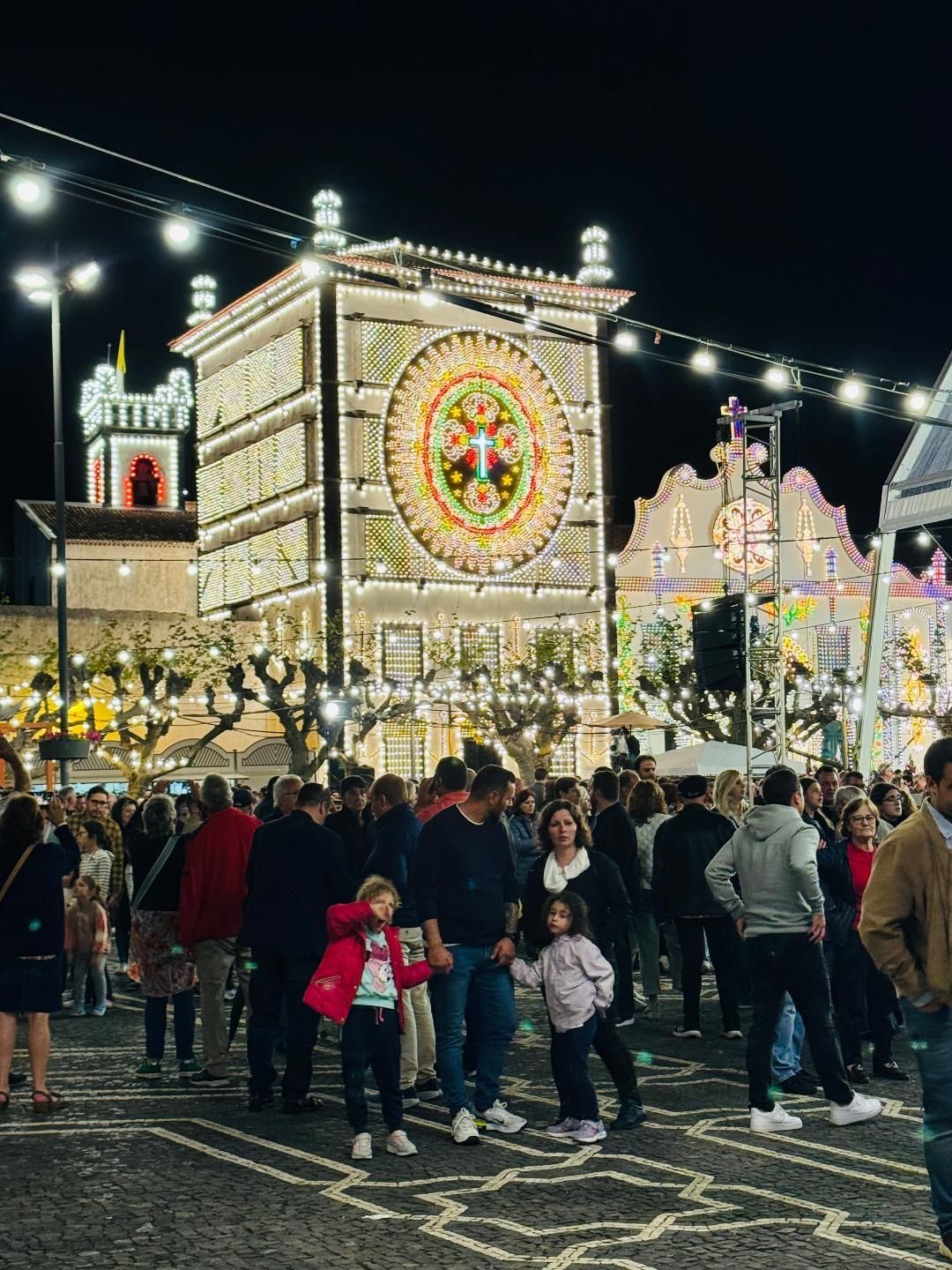 Crowd at night walking through a plaza decorated with festive, illuminated displays on buildings and overhead strings.