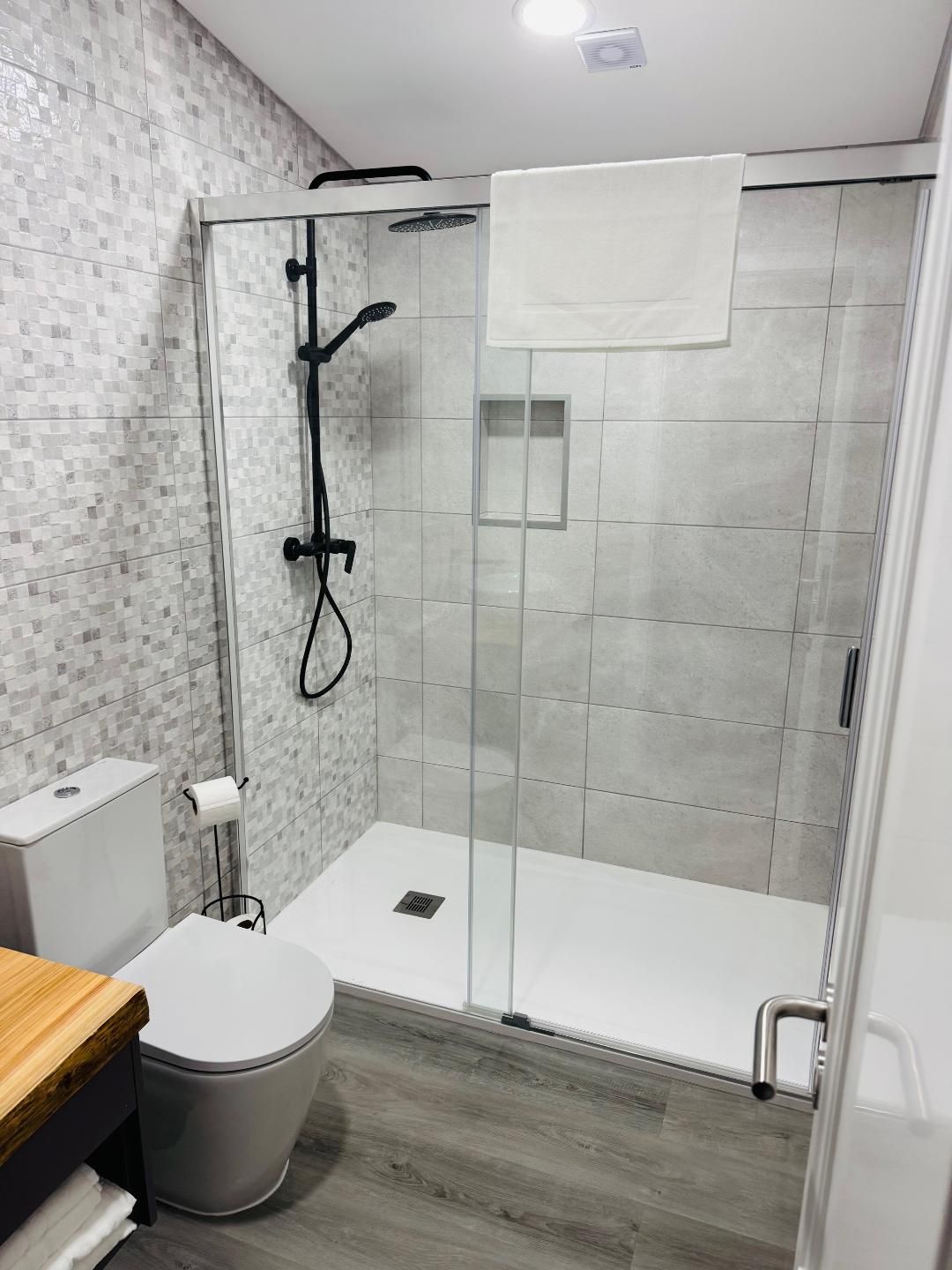 A bathroom featuring a walk-in shower with a glass door, matte black shower fixtures, a white toilet, and gray tile floors.