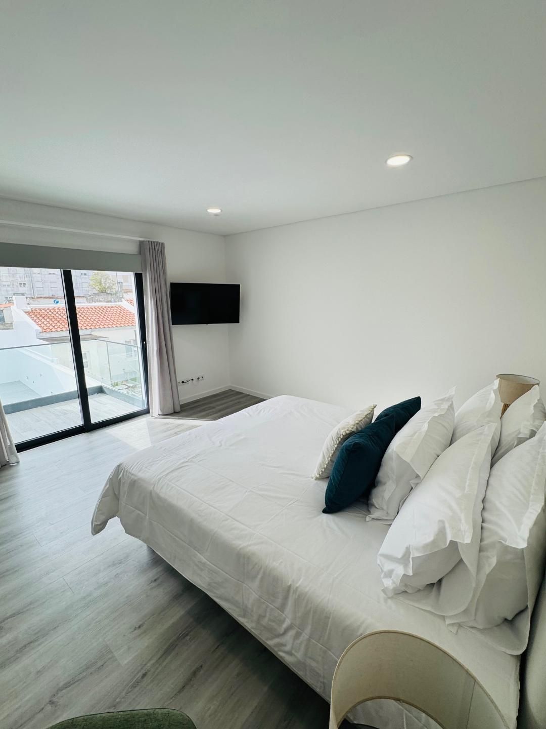 A bright, modern bedroom featuring a neatly made bed with white linens and dark pillows, beside a glass sliding door.
