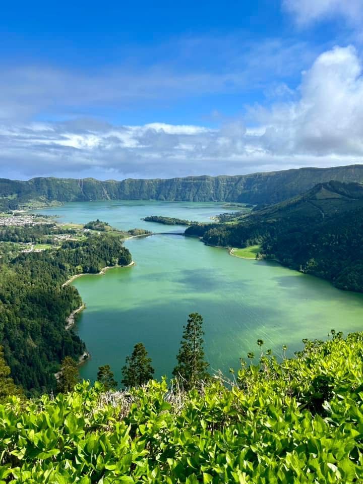A high-angle view of a large, green volcanic lake nestled within a lush, crater-rimmed landscape under a blue sky.