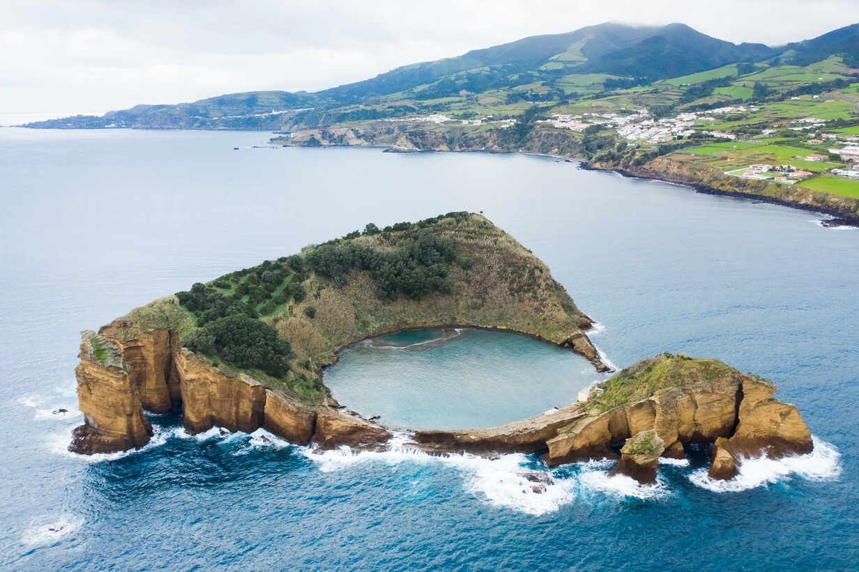 An aerial view of the Islet of Vila Franca do Campo, a circular volcanic crater filled with ocean water off a lush coast.