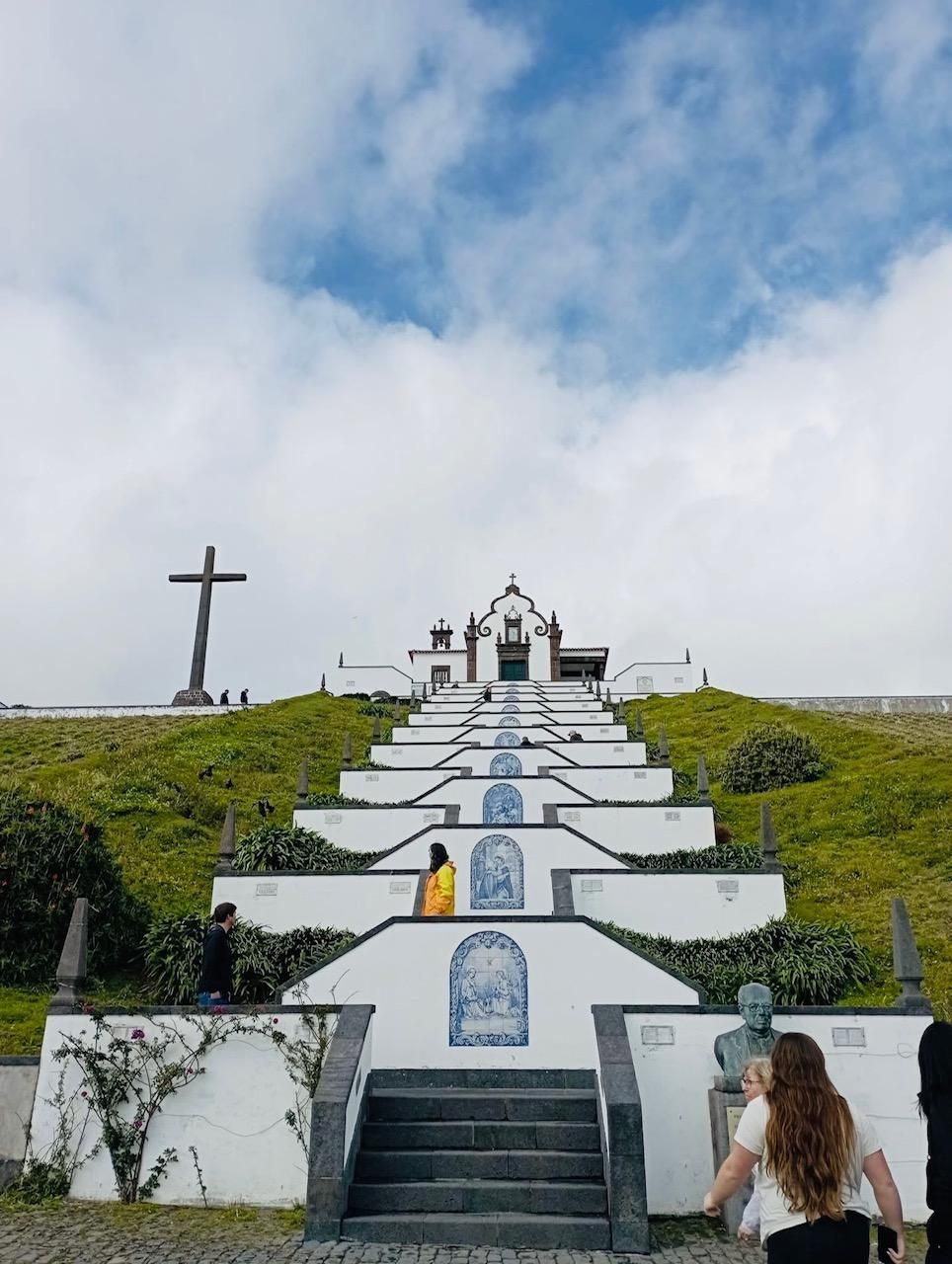 People ascend a wide, tiered stone stairway featuring tiled blue-and-white murals leading to a chapel and large cross.