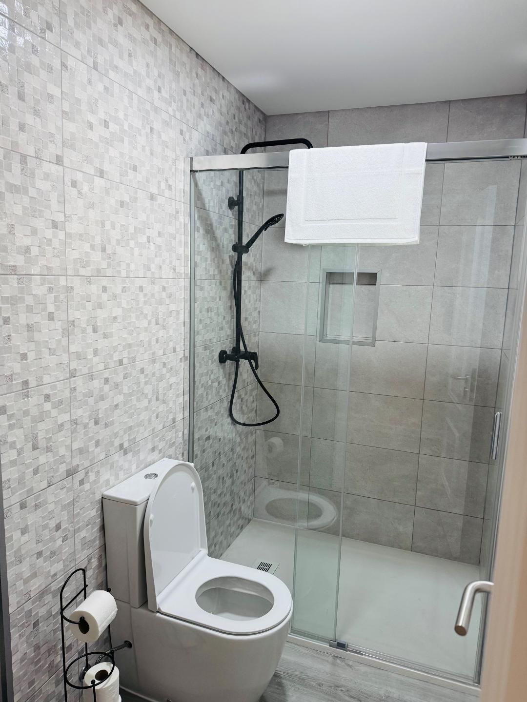 A modern bathroom featuring a white toilet, a glass-enclosed shower with black fixtures, and light grey tiled walls.
