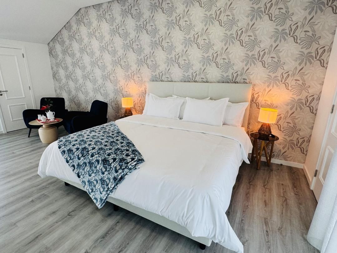 A bedroom with a large bed, patterned wallpaper, wooden flooring, two armchairs, and bedside lamps.