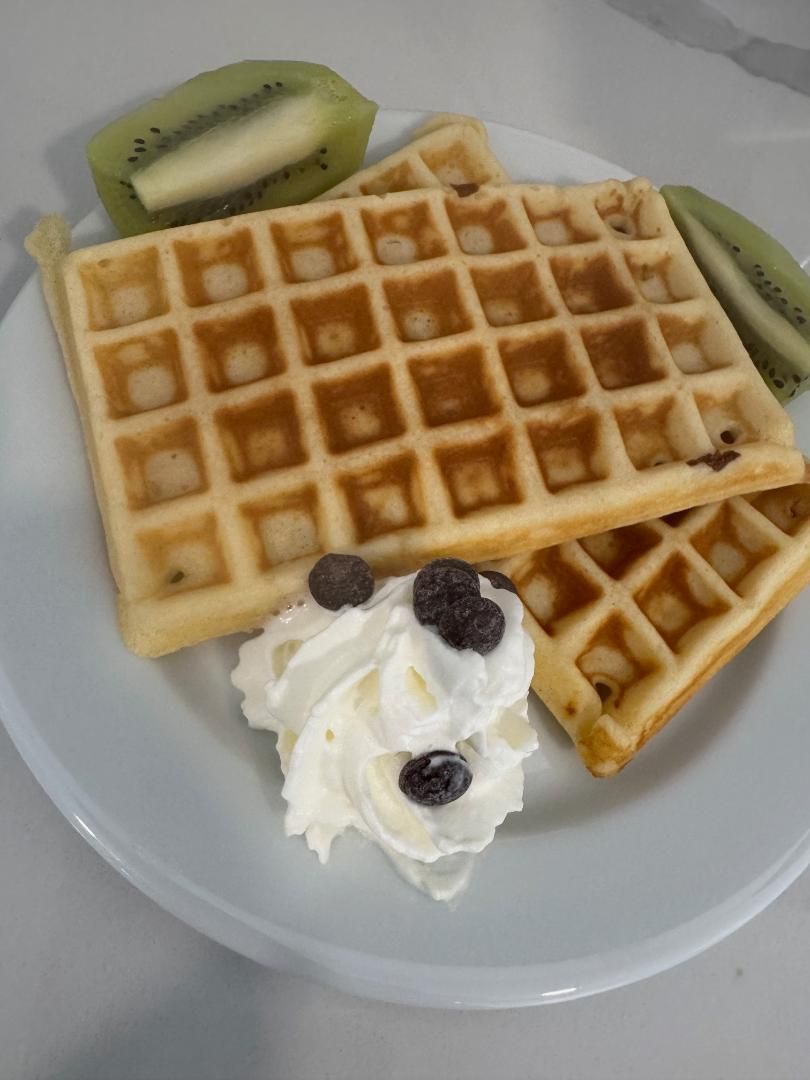 Two golden-brown waffles on a plate, topped with a dollop of whipped cream and blueberries, served with slices of kiwi.
