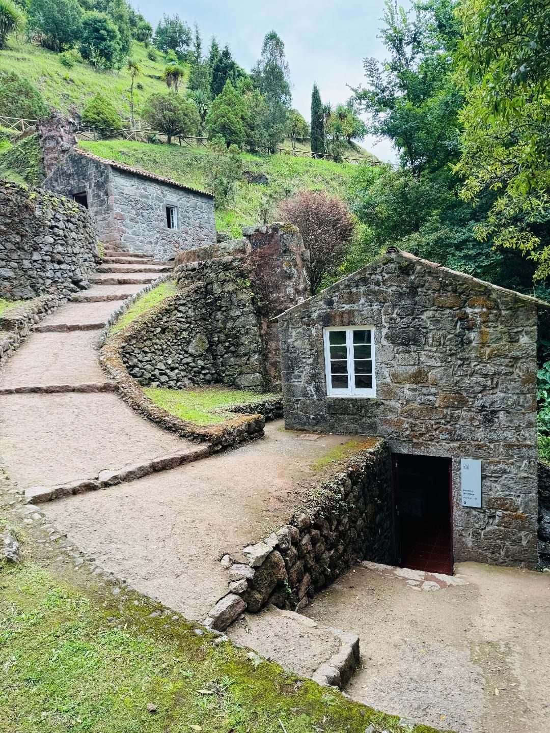 Two stone buildings sit on a grassy hillside, connected by a winding, tiered gravel path with stone retaining walls.