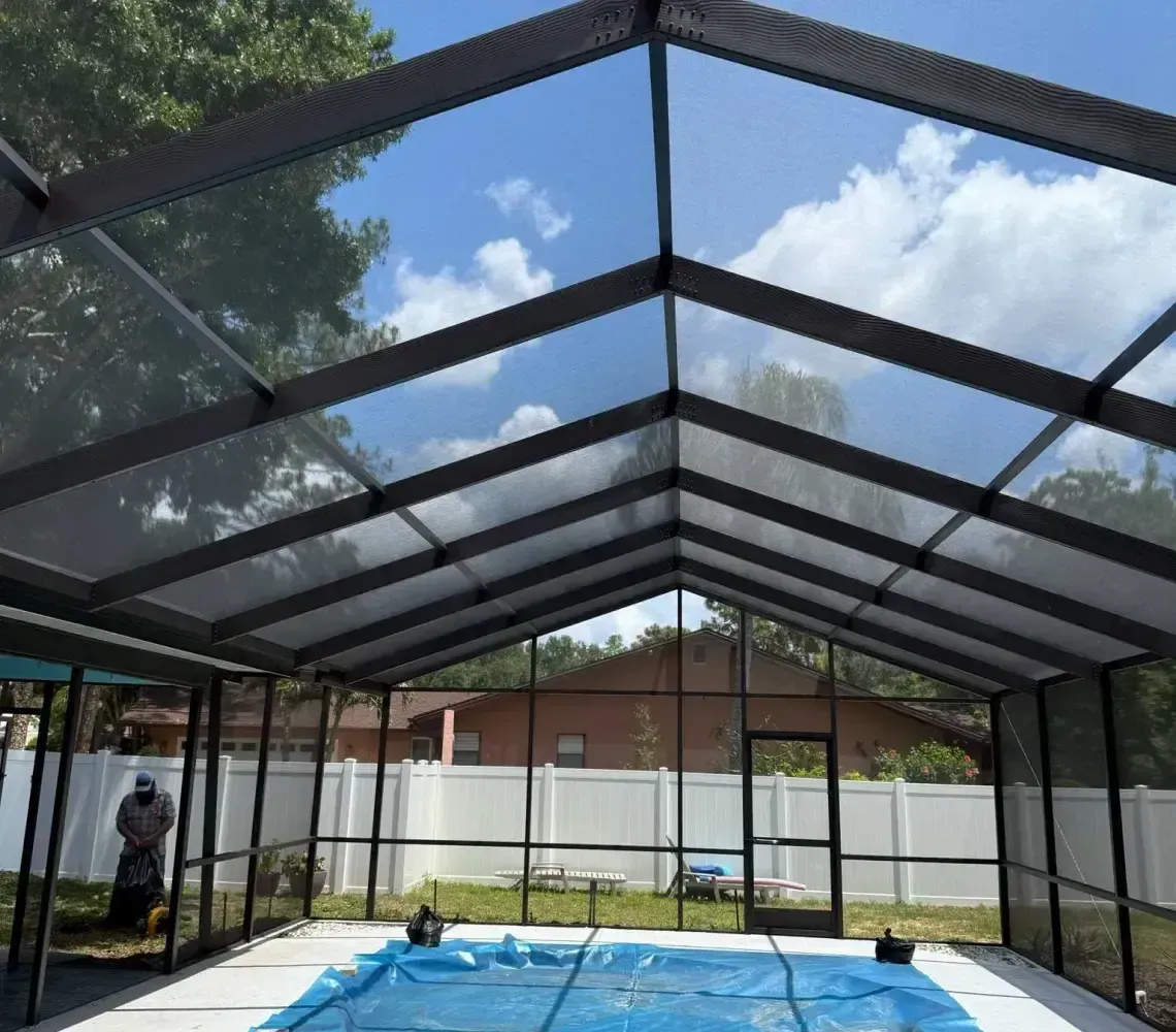 A blue pool cover lies in an enclosed pool area featuring a metal-framed screen roof under a bright, sunny sky.