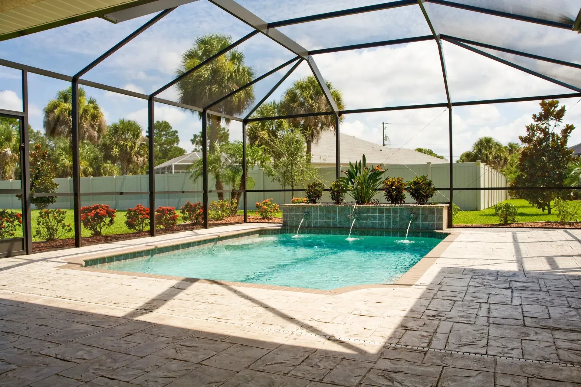 A backyard swimming pool with a fountain feature, surrounded by a stone patio and enclosed in a screened lanai.