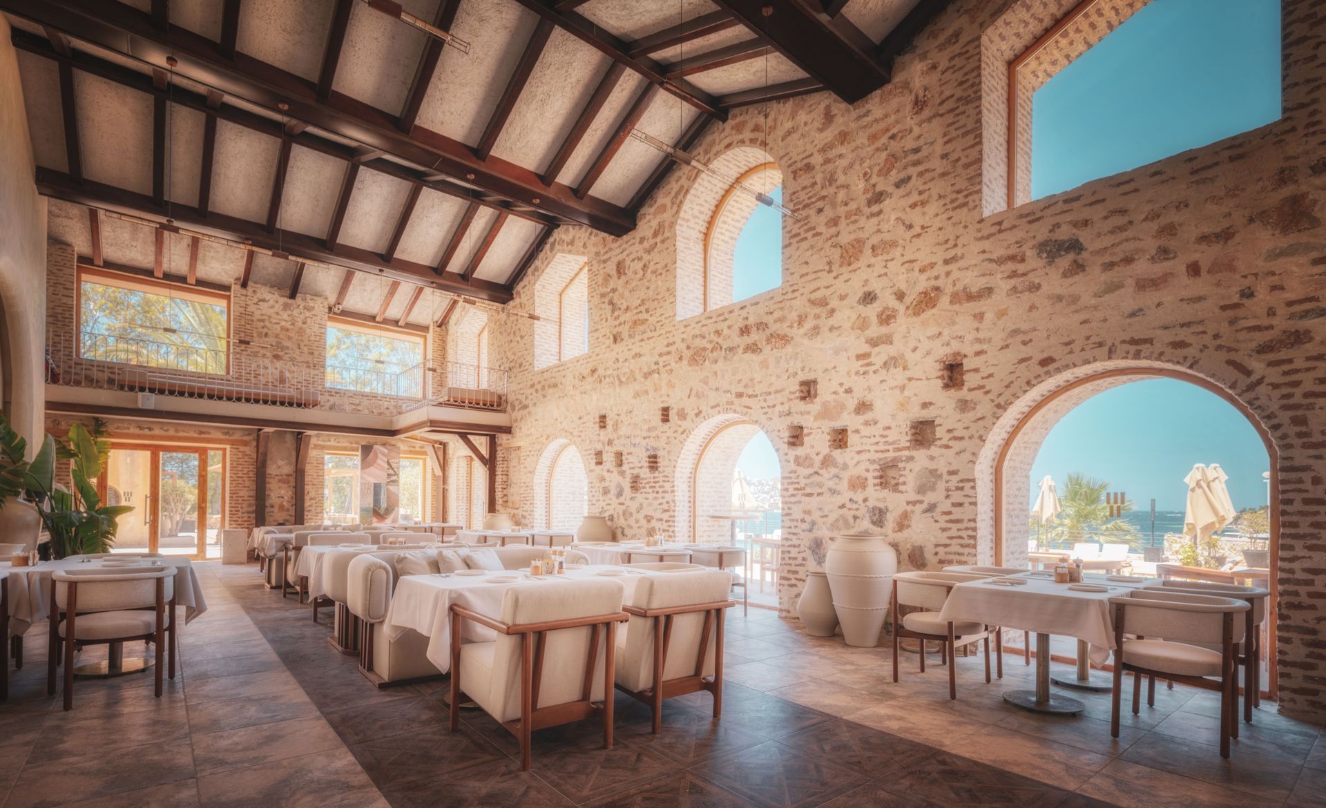Spacious rustic dining hall with stone walls, wooden beams, and white tables and chairs by arched windows