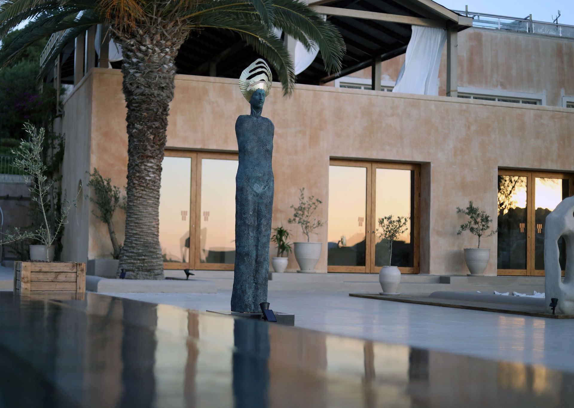 A tall, textured bronze sculpture stands in a patio before a light-colored building with large glass doors and a palm tree.