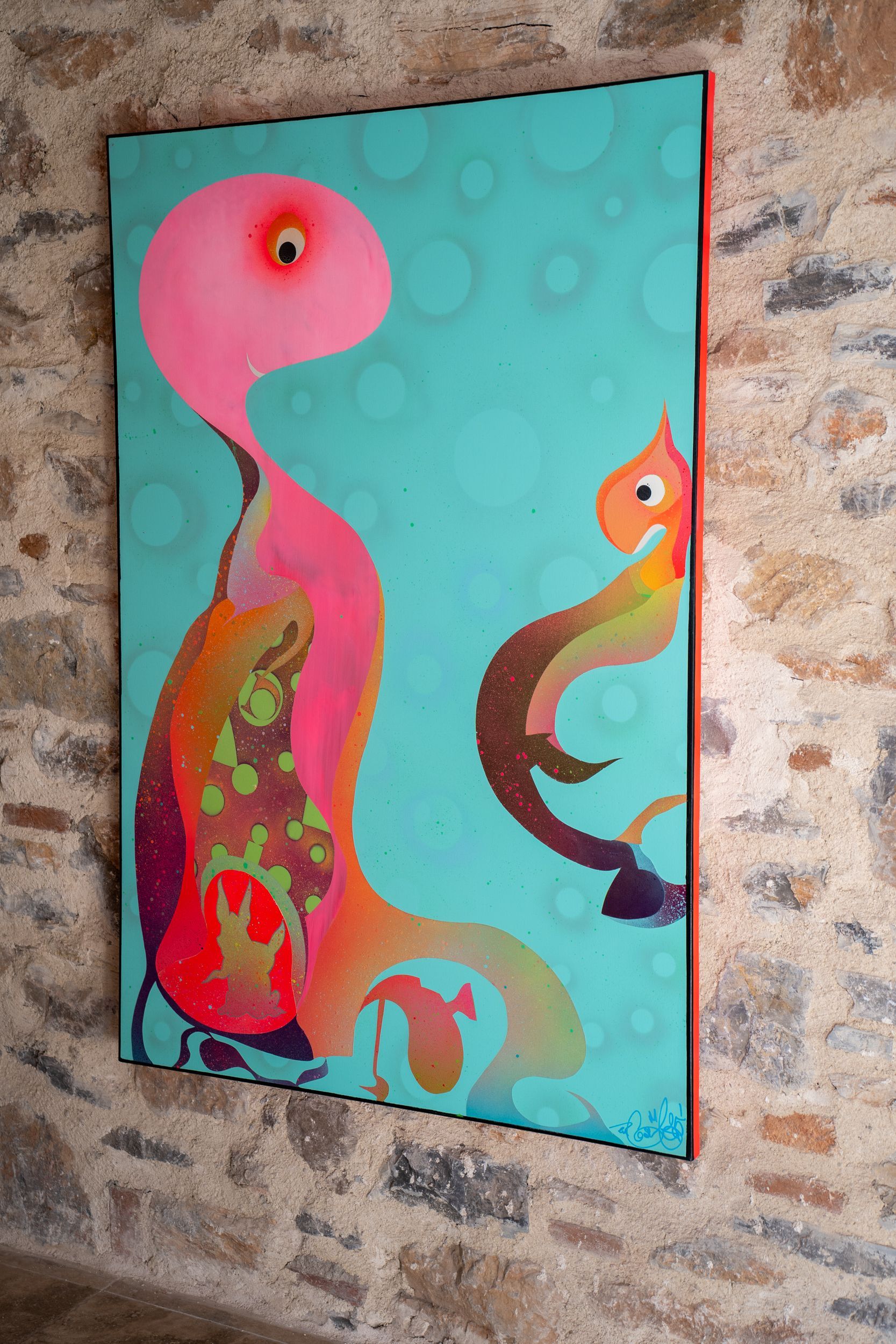 A painting of two colorful, abstract, fluid creatures floating on a turquoise background, hanging on a stone wall.