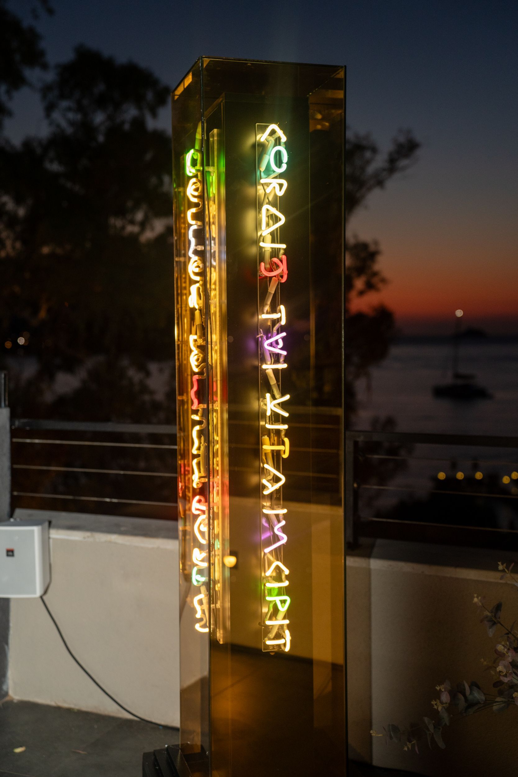A tall, glowing yellow resin pillar featuring vertical multicolored neon text at twilight near a waterfront.