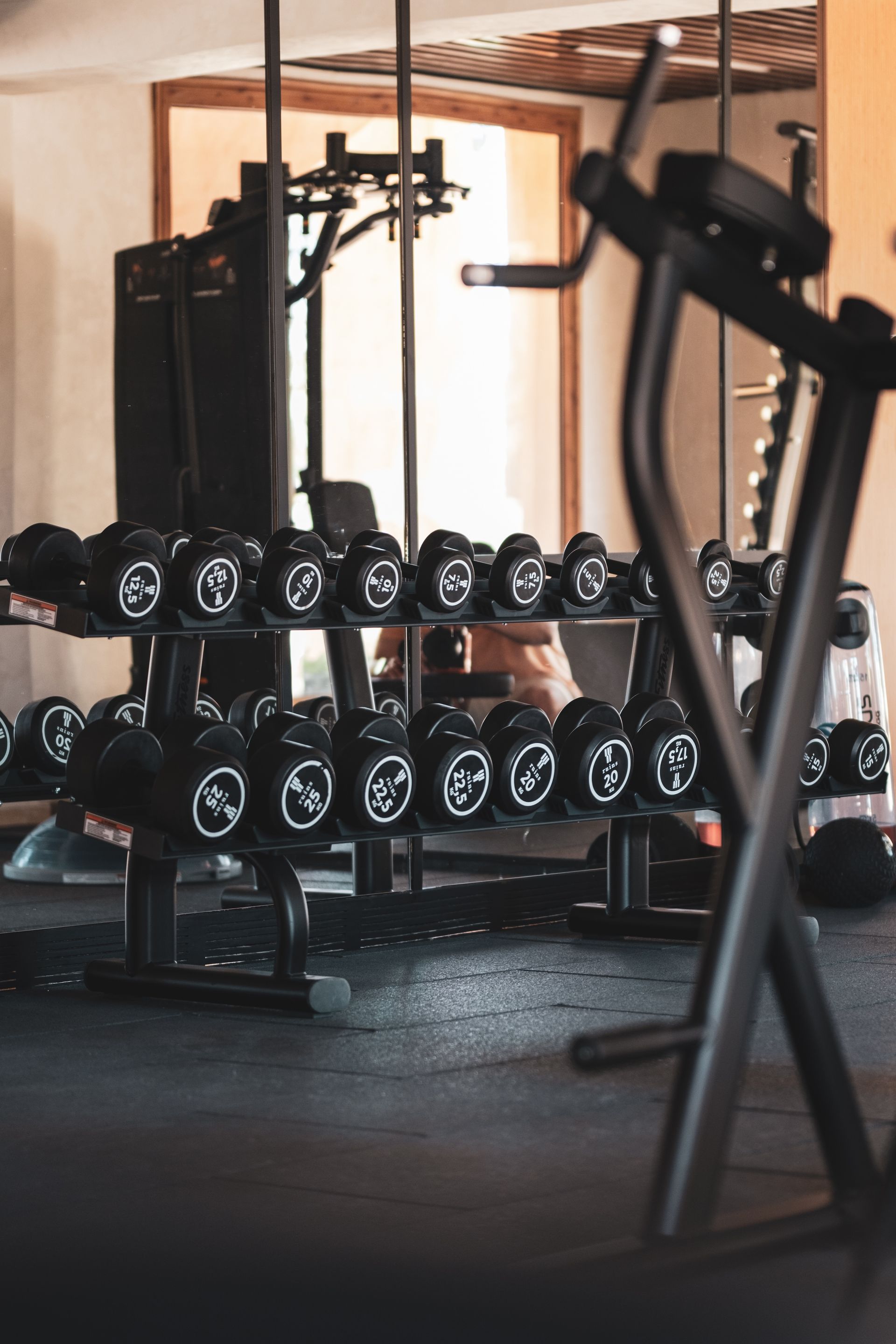A gym rack filled with rows of black dumbbells, with a portion of an exercise machine in the foreground.