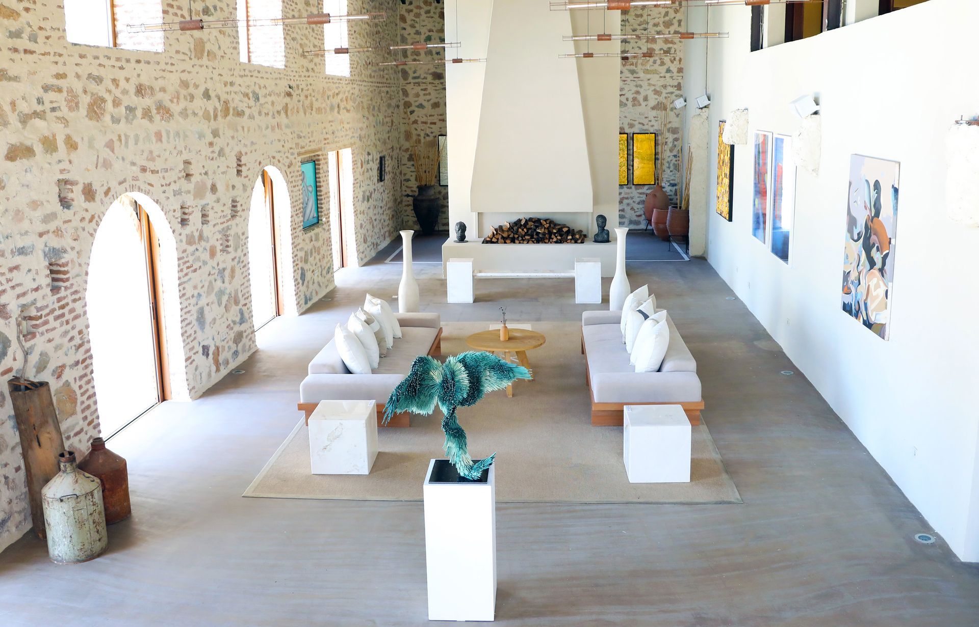 A spacious, sunlit living area with stone walls, a large central fireplace, white sofas, and a sculptural art piece.