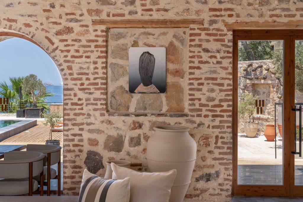 A rustic room with stone walls, an abstract framed portrait, a large ceramic jar, and an arched view of a patio.