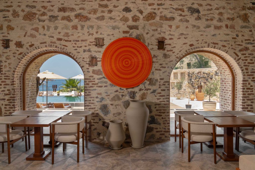 An open-air stone restaurant interior with arched windows looking out to a pool and two large minimalist floor vases.