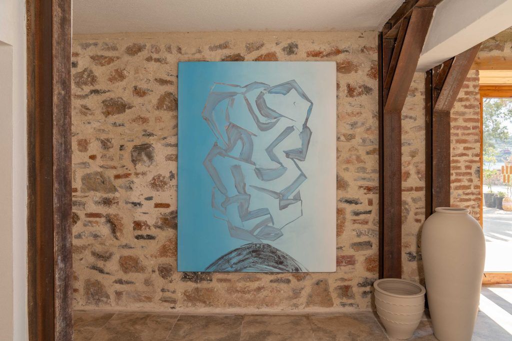 A large abstract blue painting hangs on a stone wall between wooden pillars, with two large ceramic jars in the foreground.
