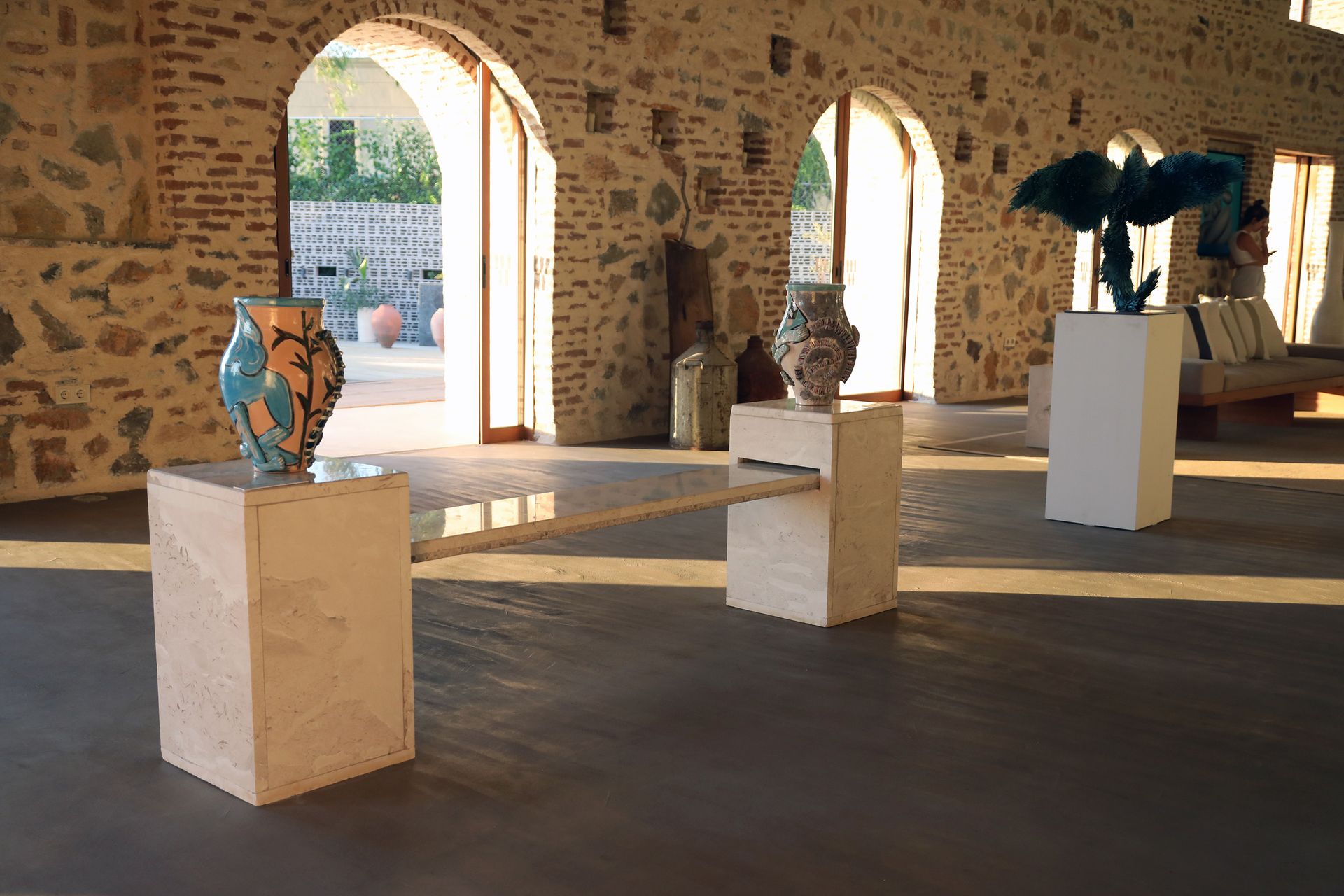 An art gallery space with stone walls and arched doors featuring sculptures displayed on stone pedestals.