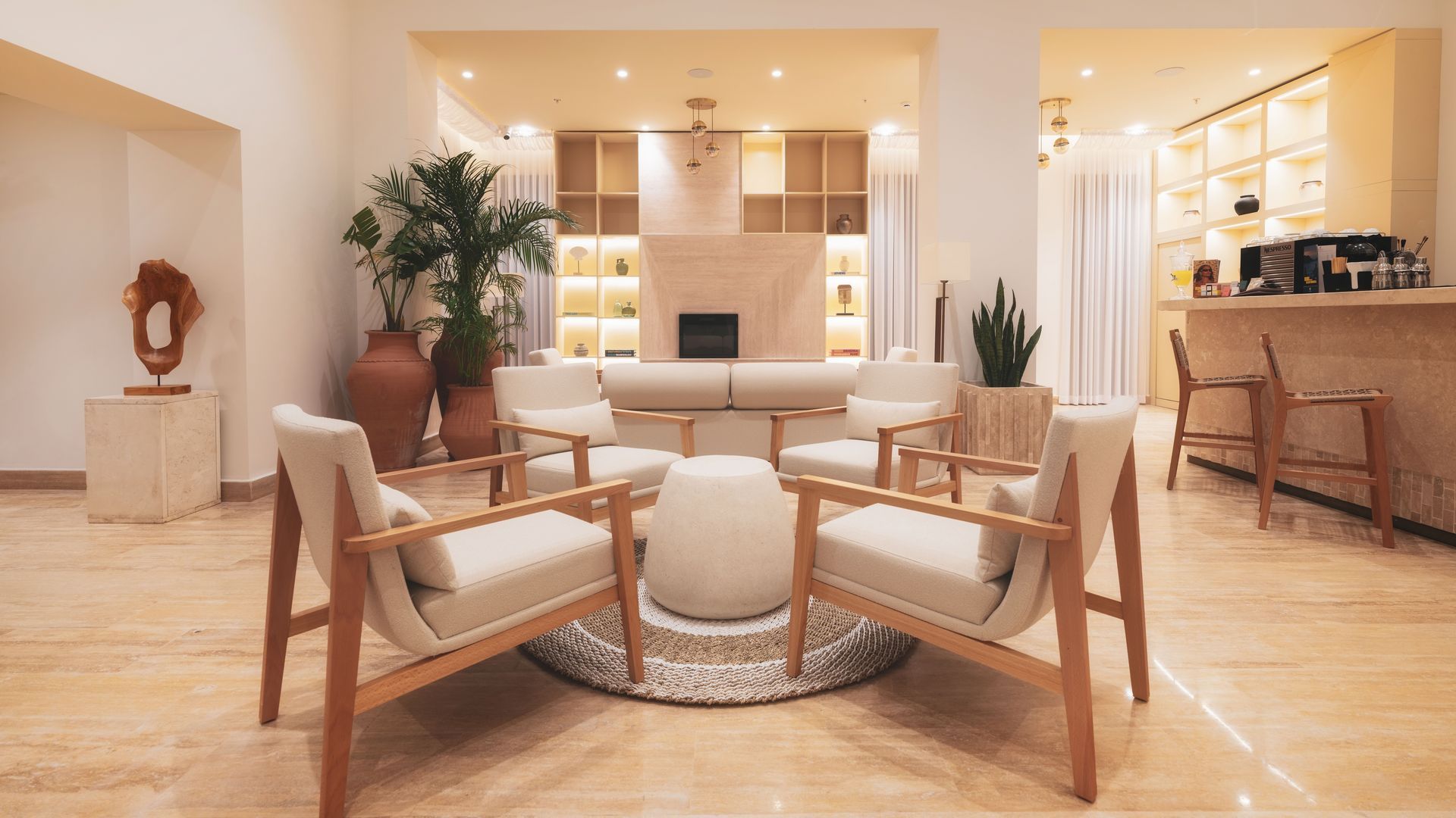 A modern, beige-toned lounge area with light wooden armchairs, a central ottoman, potted plants, and a bar area.