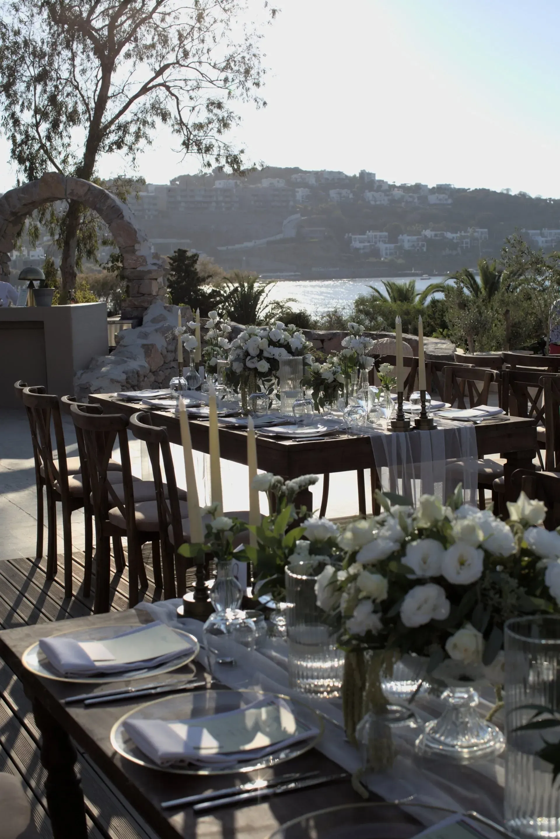 Elegant outdoor dining tables set for a wedding with white florals overlooking a sunny coastal landscape.