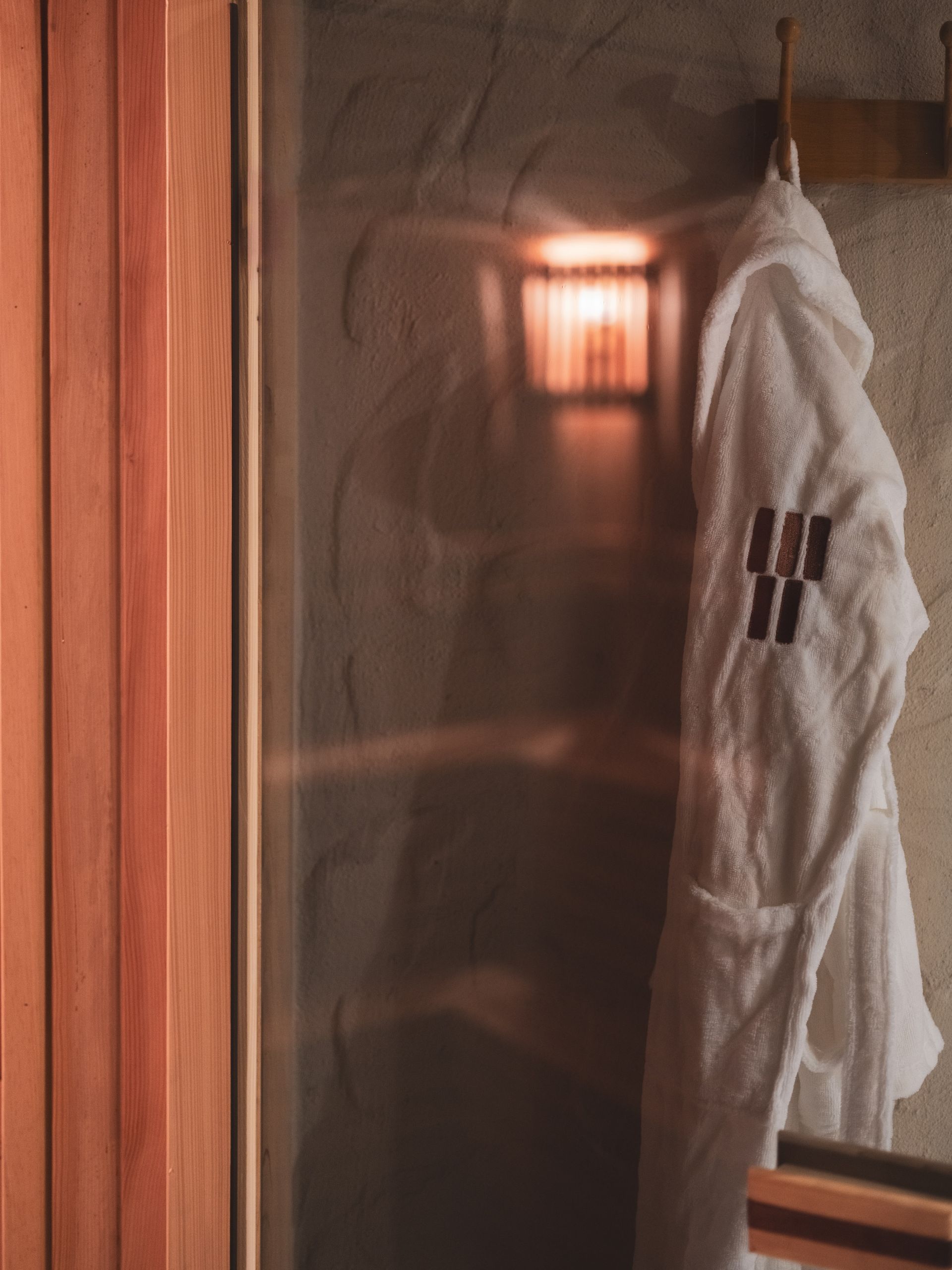 A white robe hangs on a hook next to a glass-paneled sauna door, with a glowing light visible inside.