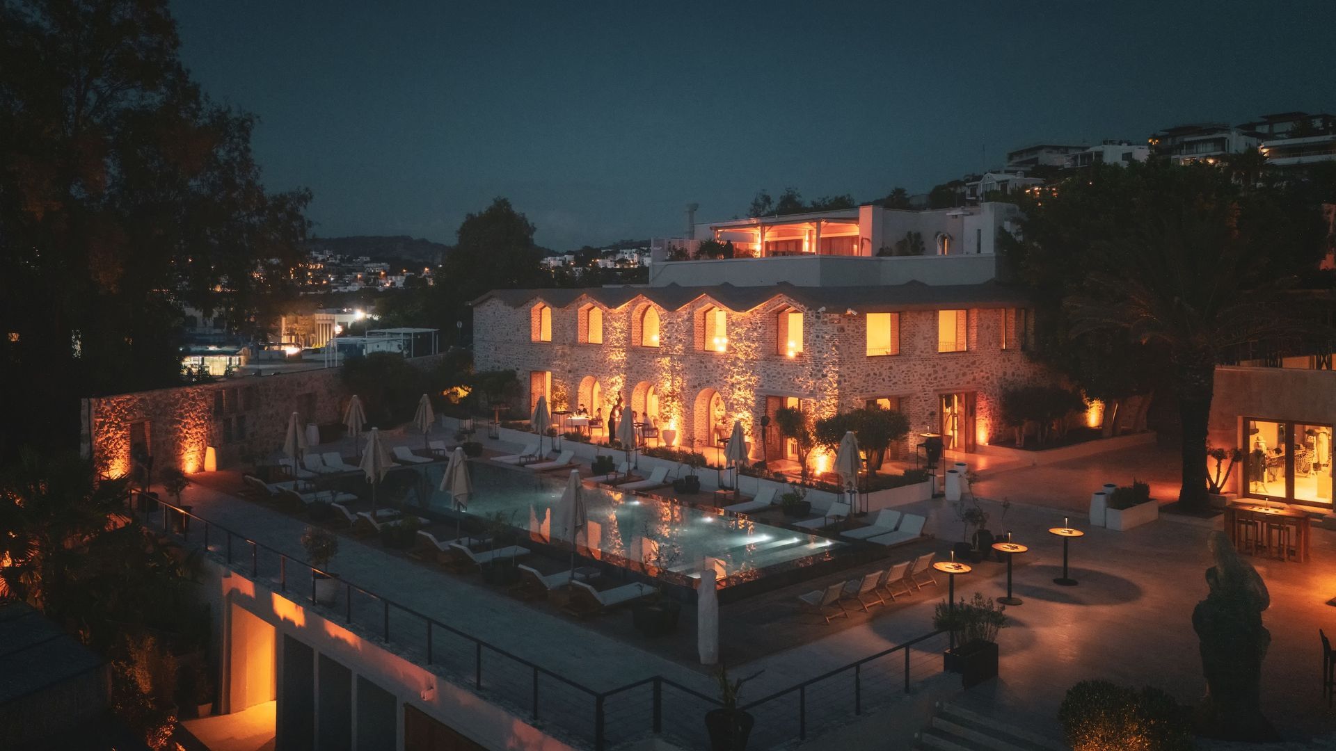 An illuminated stone hotel at night, featuring a long swimming pool surrounded by lounge chairs and warm outdoor lighting.