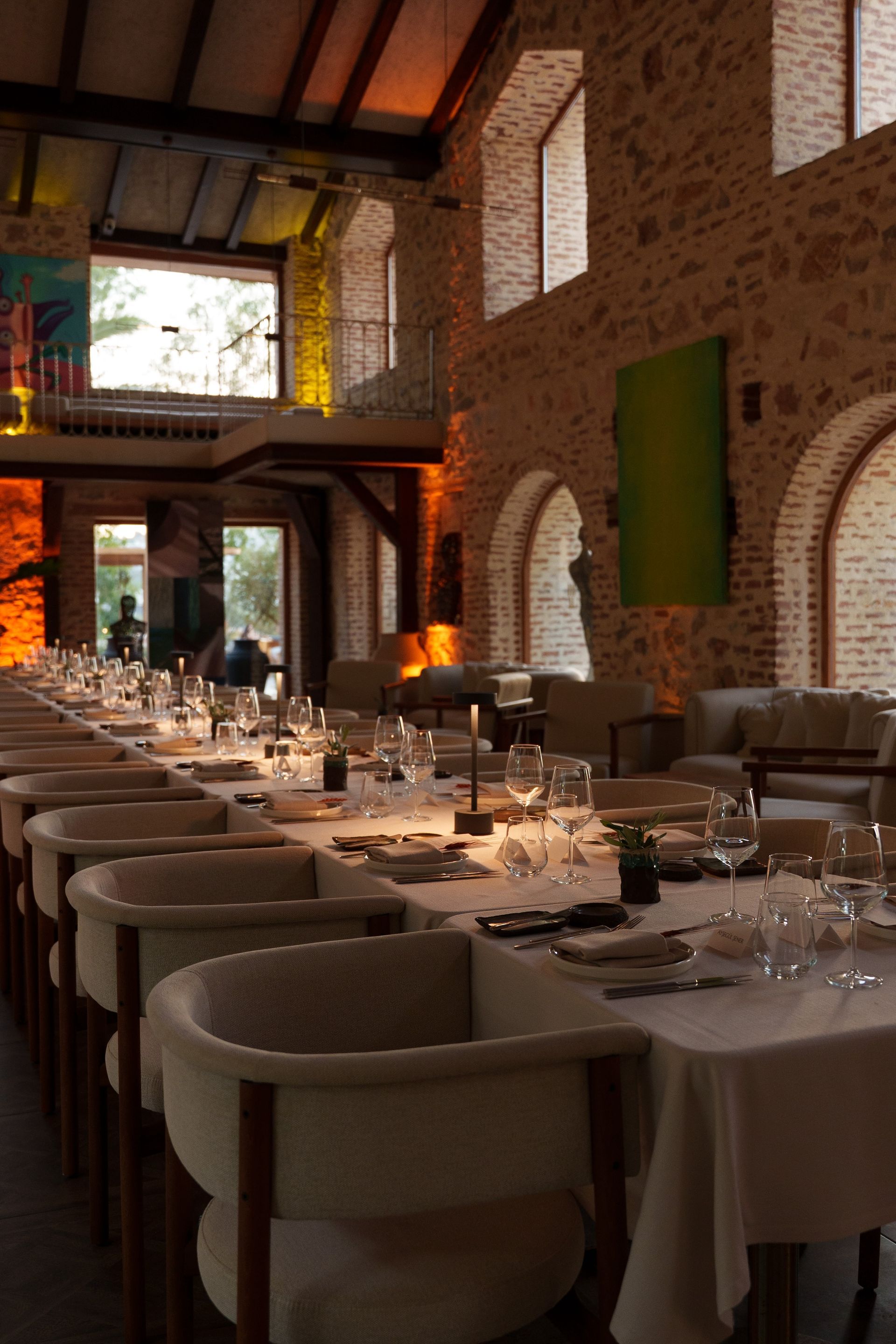 A long dining table set for an event inside a rustic stone building with arched windows and warm ambient lighting.