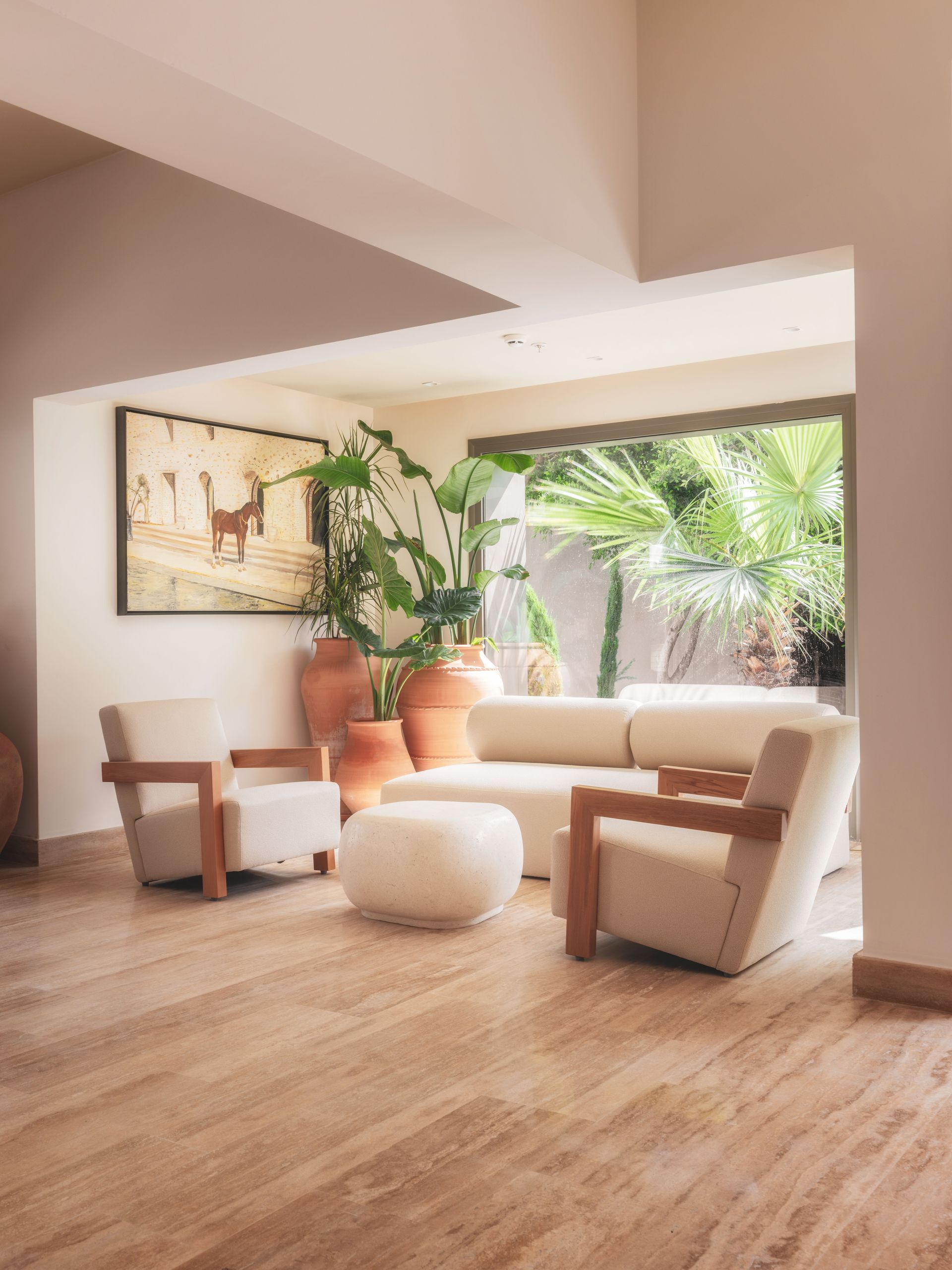 A bright, minimalist living room with a beige sofa, two wooden chairs, a white ottoman, and large potted plants.