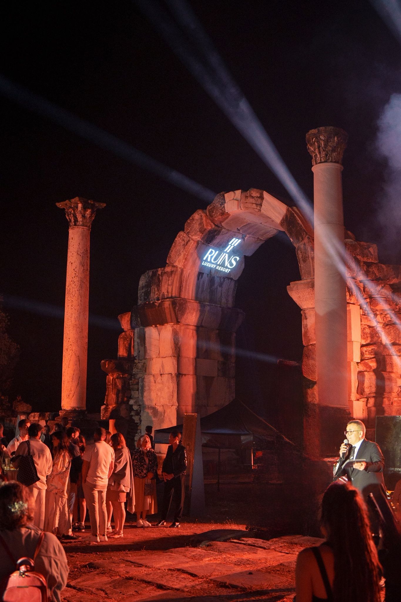 A performer speaks on a stage with dramatic spotlights and light projections against ancient stone ruins at night.