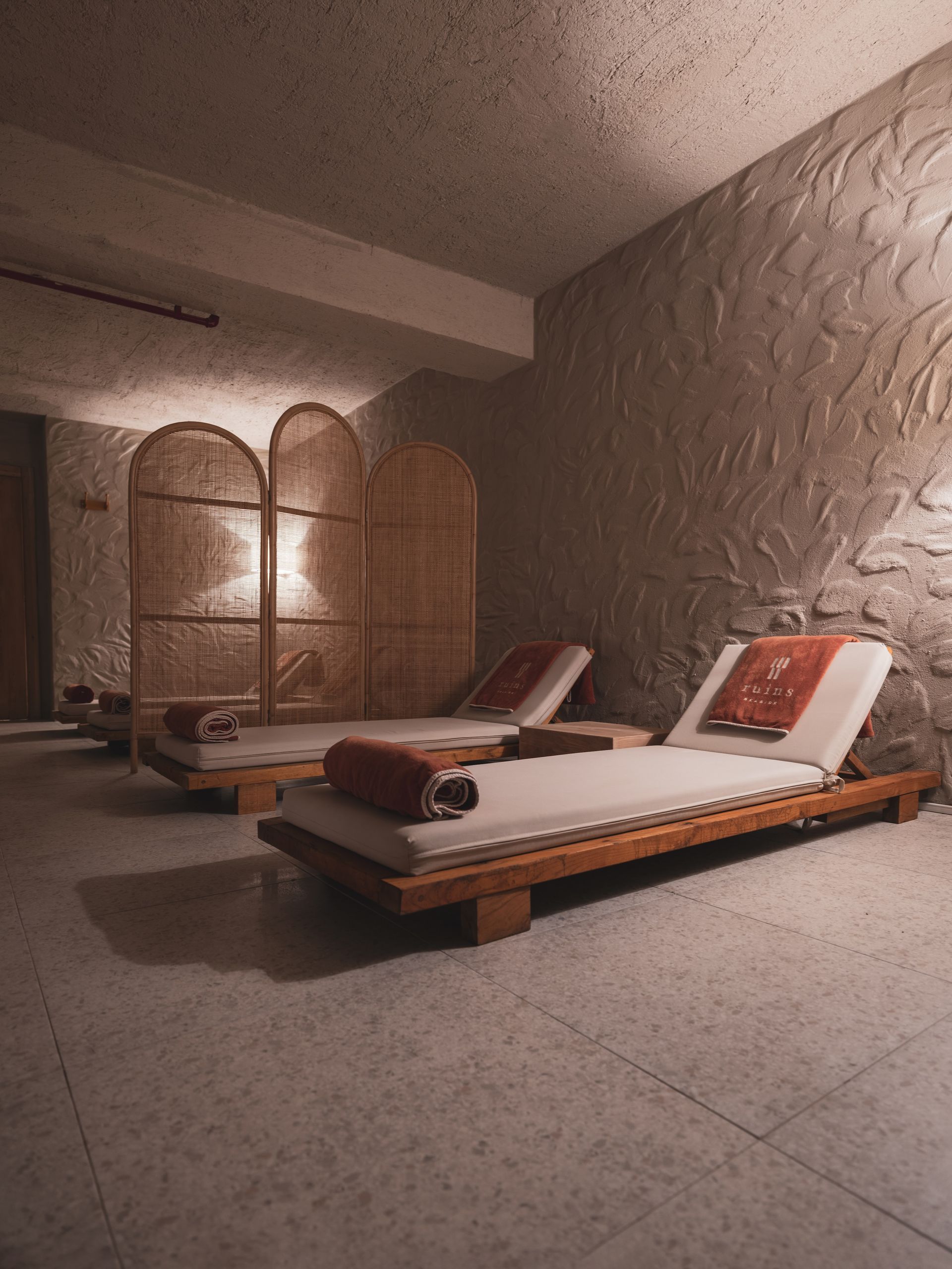 Two wooden lounge chairs with white cushions and red accents in a dimly lit, stone-walled spa room with a wicker screen.