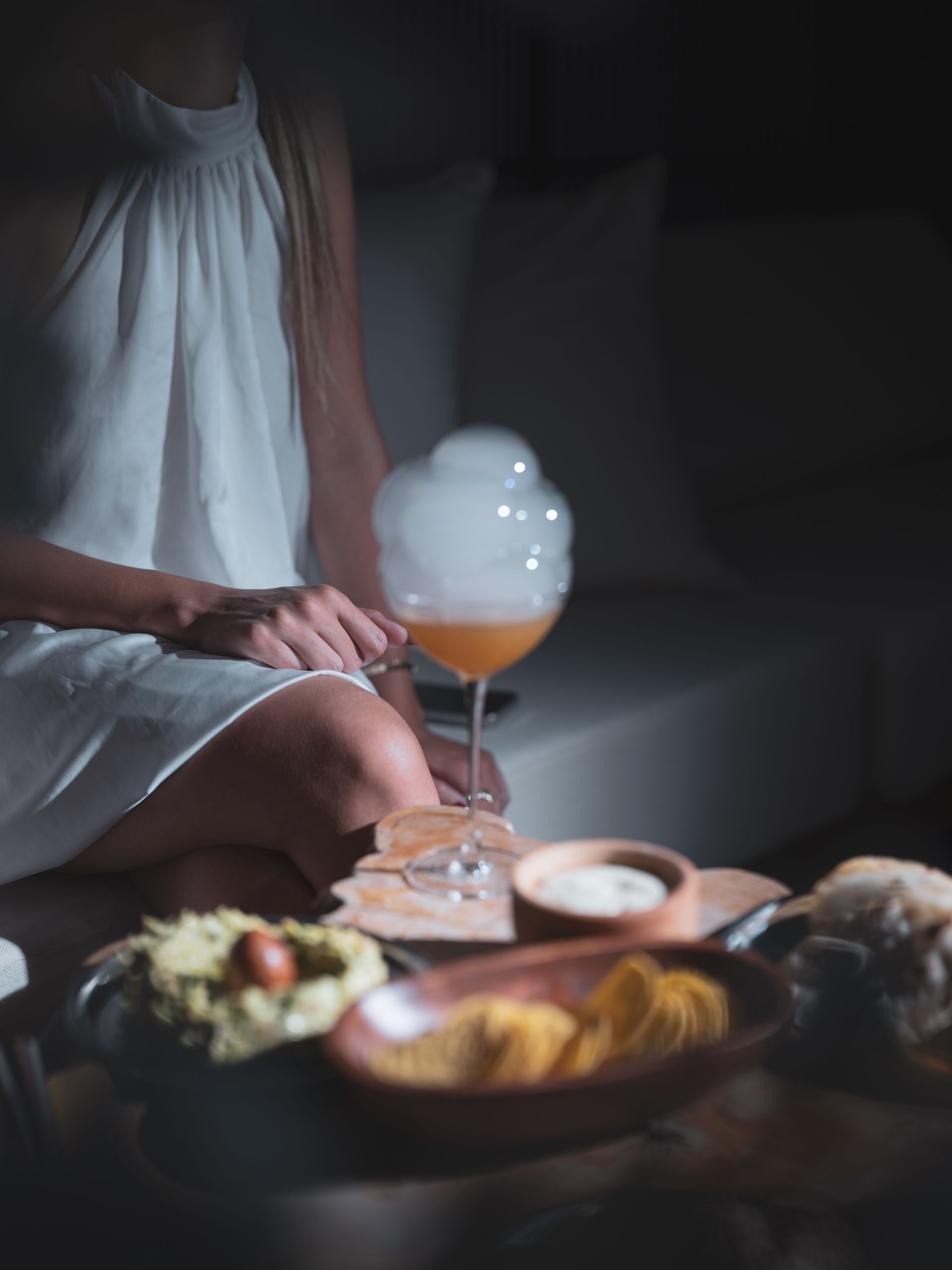 A person in a white dress sits at a table with a smoking cocktail and bowls of food in a dim, modern lounge setting.