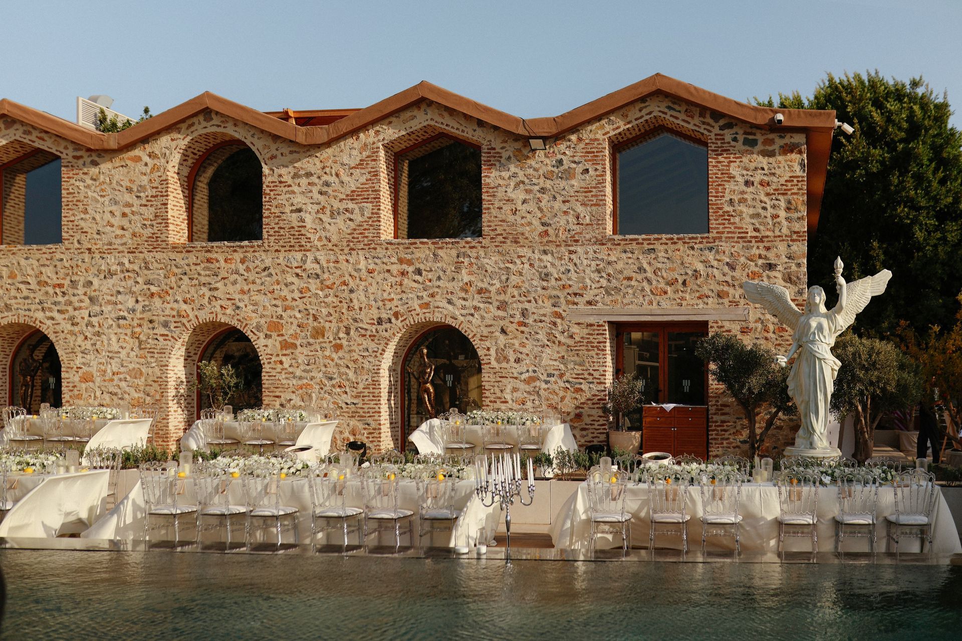 An outdoor dining set-up with white chairs and tables beside a pool in front of a rustic, stone-walled building.