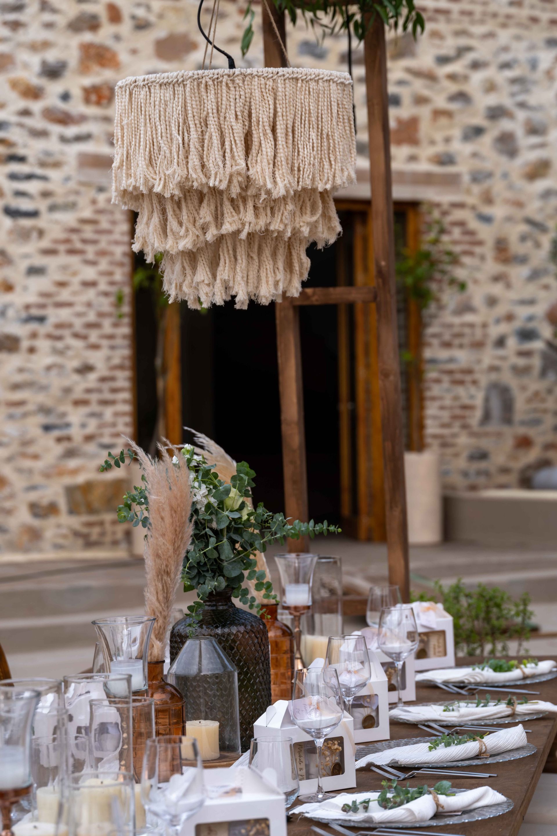 A bohemian-style table setting for an event, featuring a macramé chandelier, greenery, and dried pampas grass.
