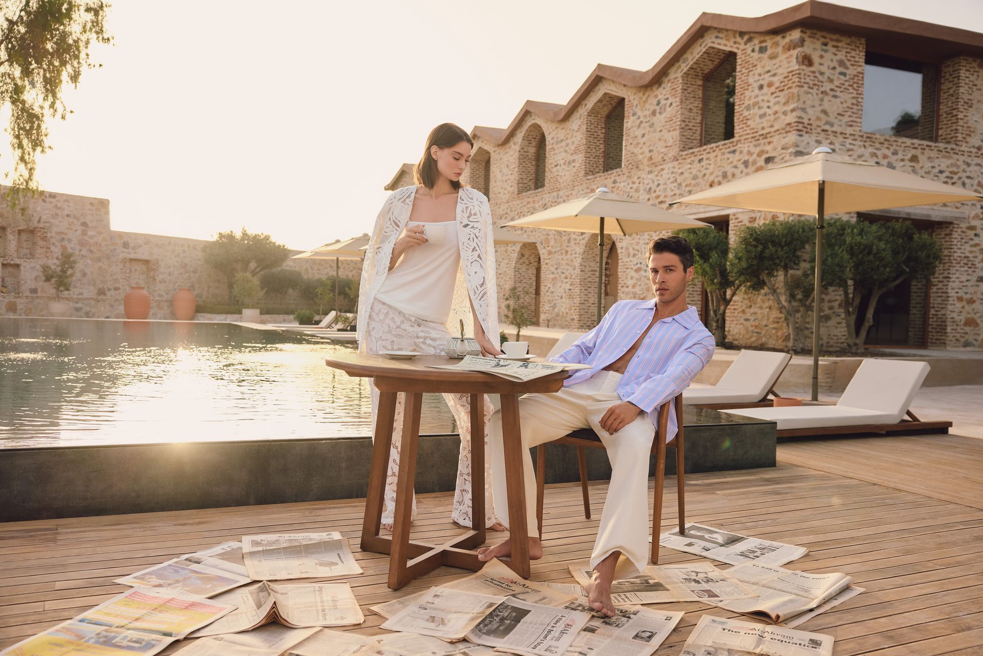 A person standing and another seated at a wooden table outdoors, with scattered newspapers on the ground by a pool.