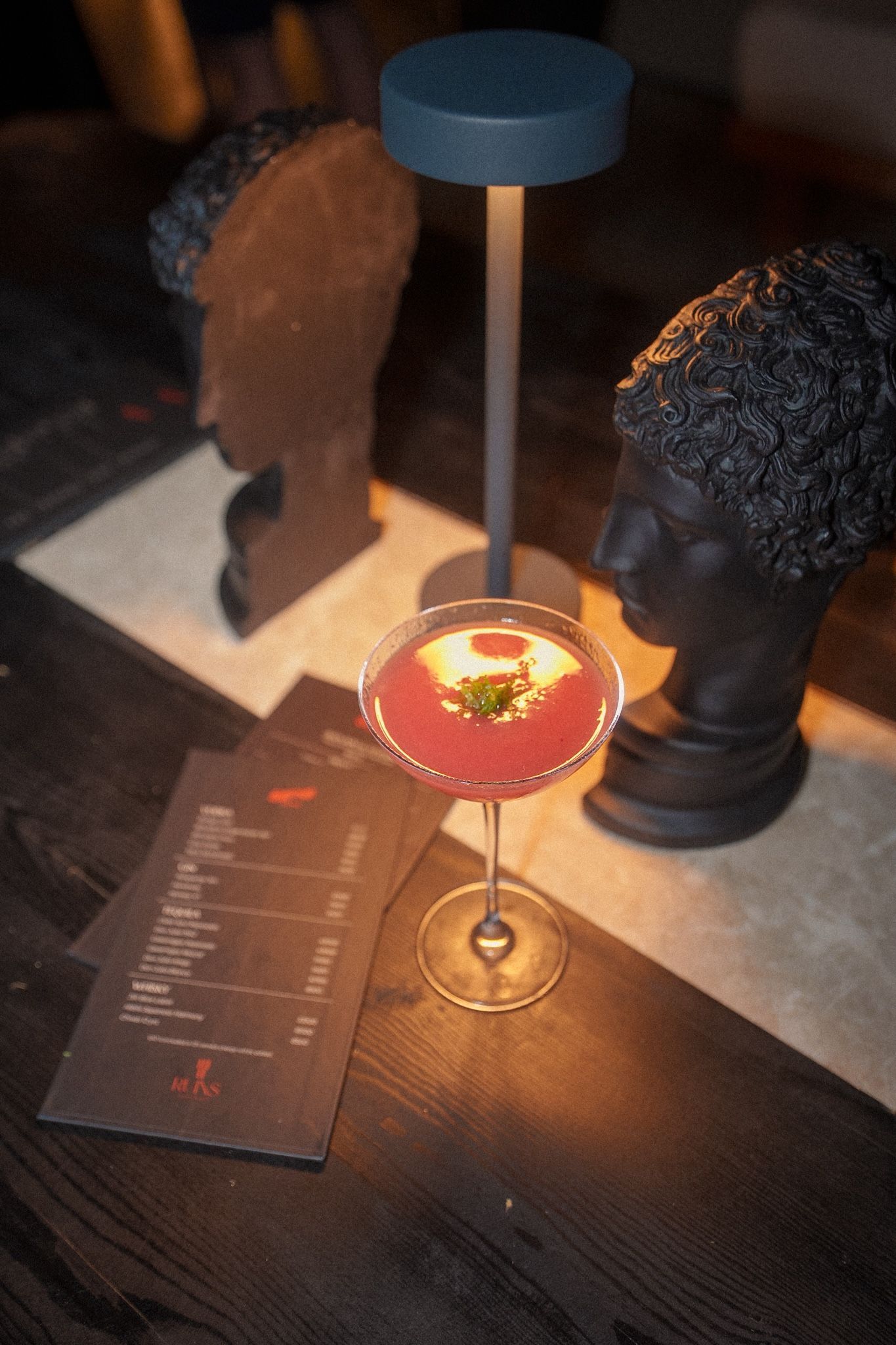 A pink cocktail in a stemmed glass sits on a wooden table next to a menu and two decorative sculpted heads.