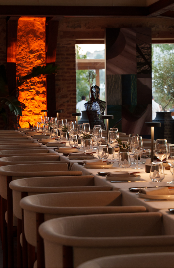 A long, elegantly set dinner table with modern armchairs in a dimly lit, upscale restaurant with warm accent lighting.