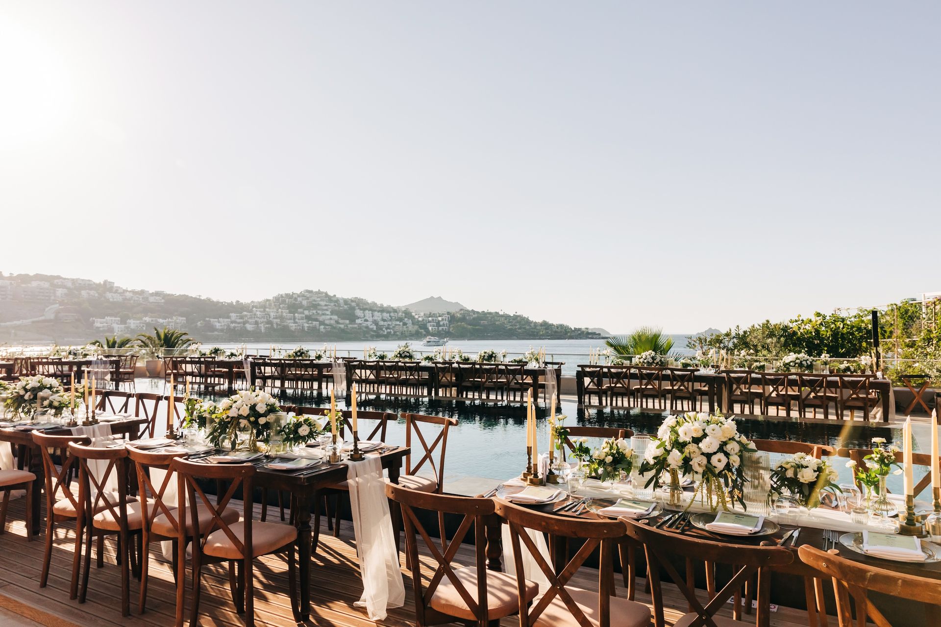 An outdoor wedding reception with wooden tables and chairs set by the sea, featuring white floral arrangements.