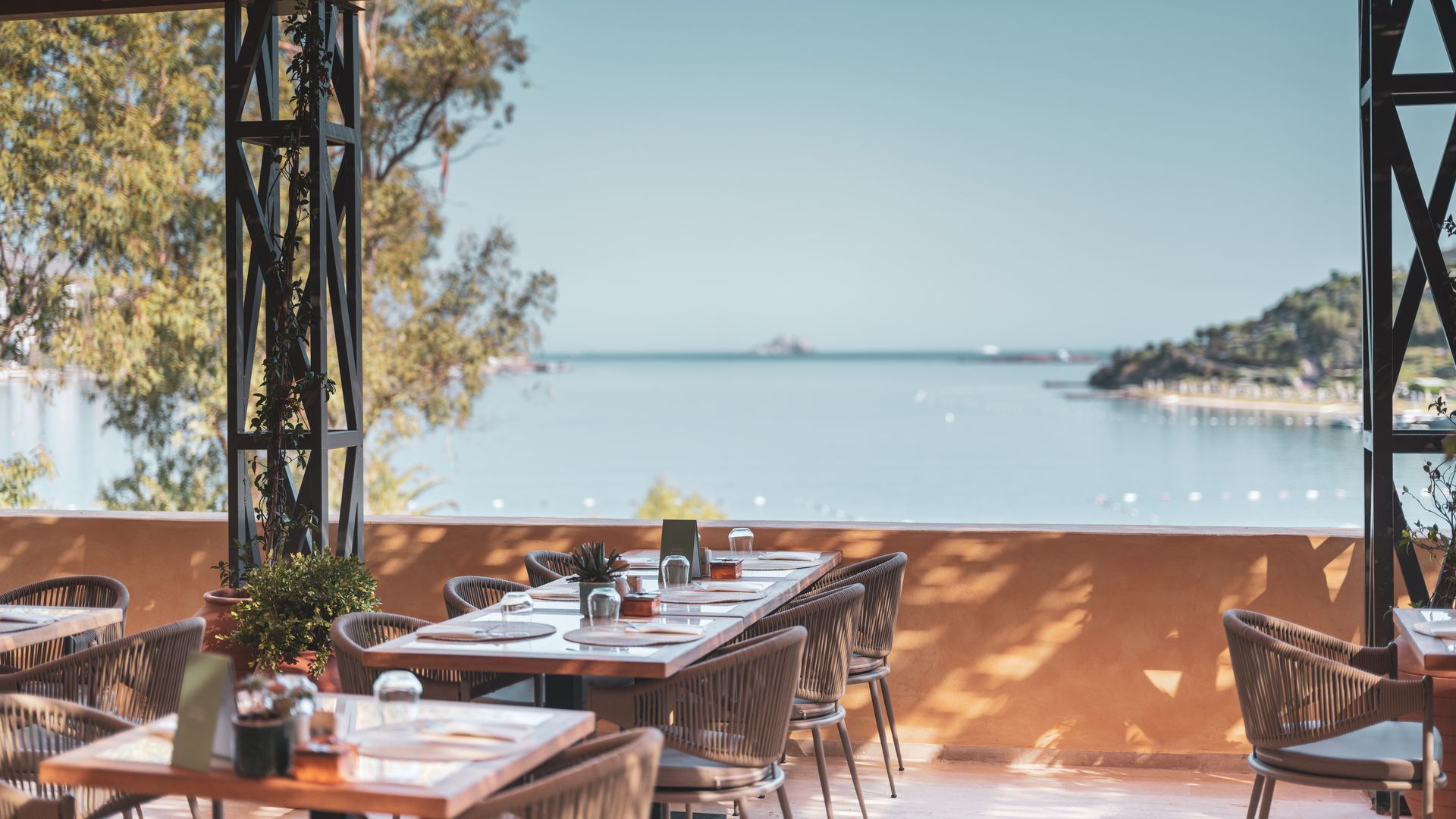 An open-air restaurant patio overlooking a calm bay with trees, sand, and blue water under a clear sky.